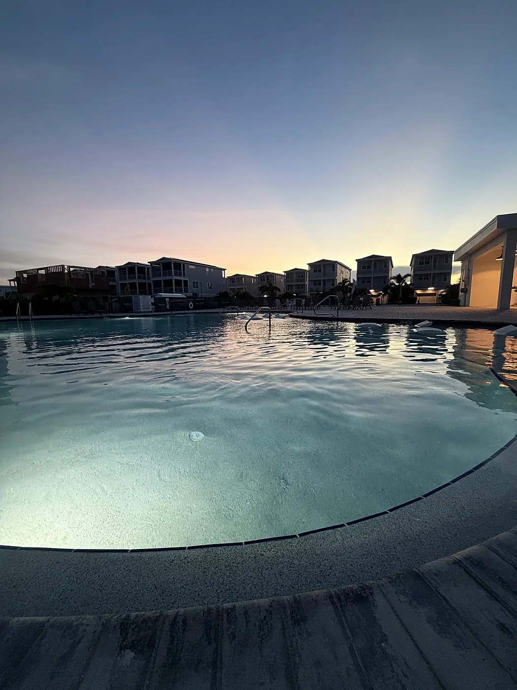 Community pool lit from below at dusk, with townhomes and a pastel sunset behind it